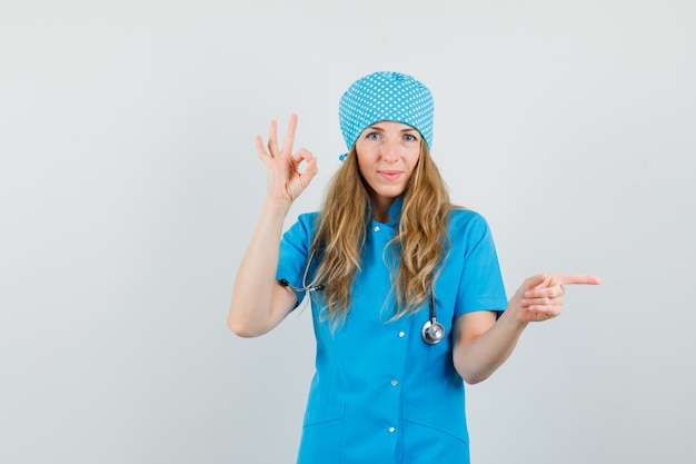 Ärztin in blauer Uniform zeigt OK-Zeichen, während sie zur Seite zeigt und fröhlich aussieht