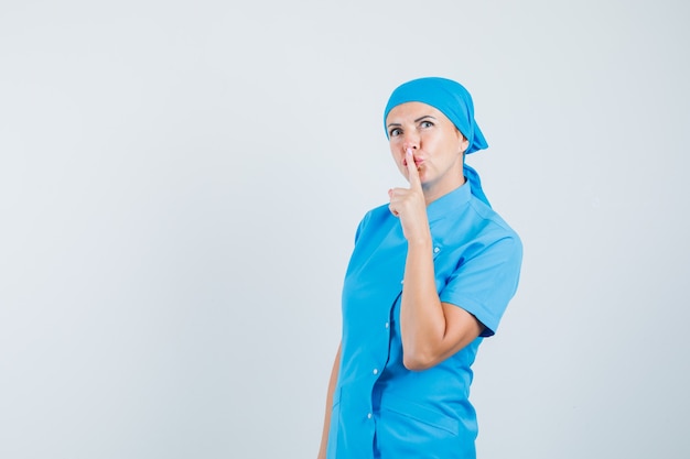 Ärztin in blauer Uniform zeigt Schweigegeste und schaut vorsichtig, Vorderansicht.