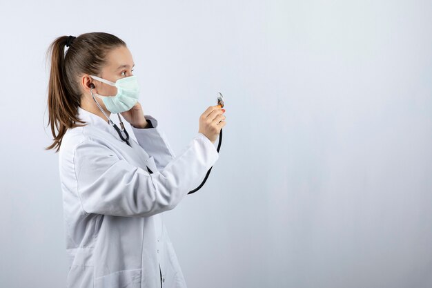 Ärztin in weißer Uniform, die eine medizinische Maske trägt und ein Stethoskop hält