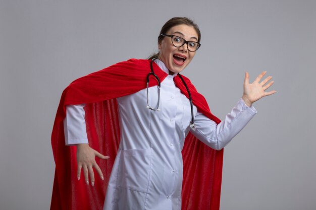 Ärztin in weißer Uniform und rotem Superheldenumhang, der überrascht und glücklich aussieht, bereit zu helfen, über weißer Wand zu stehen
