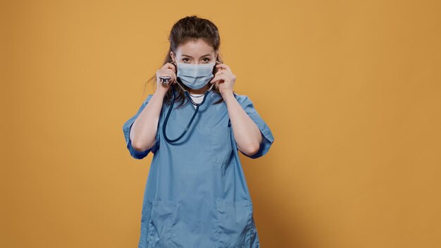 Ärztin mit Covid-Maske zum Schutz vor Viren, Stethoskop aufsetzen und im Studio vor der Kamera zeigen. Porträt eines kaukasischen Sanitäters mit Schutzuniform und medizinischem Gerät.