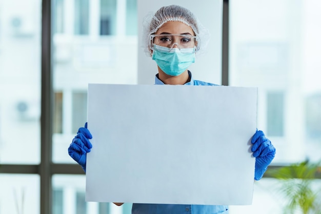 Ärztin mit Schutzkleidung, die leere Pappe hält, während sie in der Klinik steht und in die Kamera schaut Kopierraum