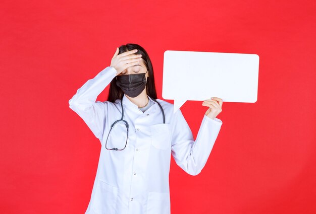Ärztin mit Stethoskop und in schwarzer Maske, die einen rechteckigen Infotisch hält und Kopf hält