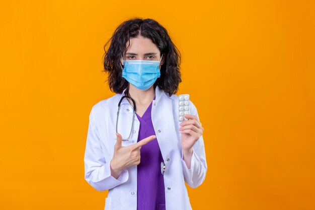 Ärztin mit weißem Mantel und Stethoskop in der medizinischen Schutzmaske, die mit dem Finger auf die Blase mit Pillen mit ernstem Gesicht auf isoliertem Orange zeigt