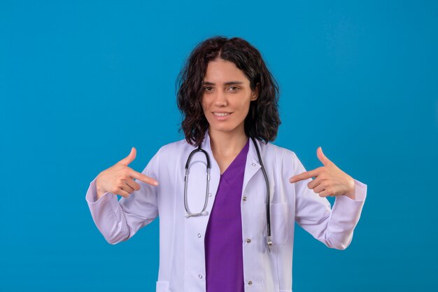 Ärztin mit weißem Mantel und Stethoskop zeigt auf sich selbst mit Daumen, die auf isoliertem Blau zuversichtlich aussehen