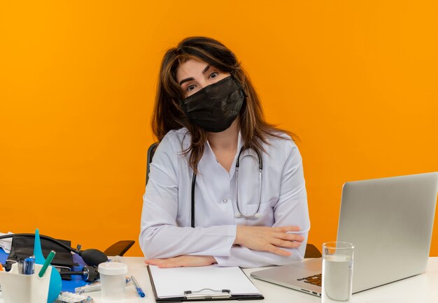 Ärztin mittleren Alters, die medizinische Robe mit Stethoskop in der medizinischen Maske trägt, die am Schreibtisch sitzt, arbeiten am Laptop mit medizinischen Werkzeugen auf isolierter orange Wand mit Kopienraum