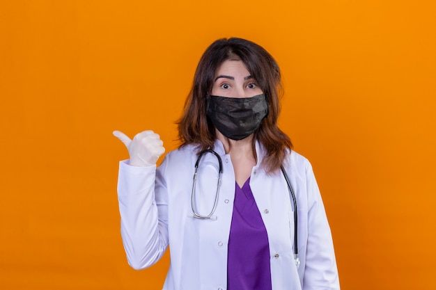 Ärztin mittleren Alters, die weißen Kittel in schwarzer schützender Gesichtsmaske trägt und mit Stethoskop betrachtet Kamera überrascht zeigt, zeigt auf die Seite mit Daumen nach oben stehend über isoliertem orangefarbenem Bac