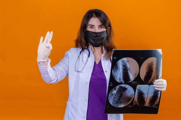 Ärztin mittleren Alters, die weißen Kittel in schwarzer schützender Gesichtsmaske trägt und mit Stethoskop hält Röntgenaufnahme der Lunge, die Kamera positiv tut, das ok Zeichen steht über orange Hintergrund