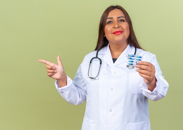 Ärztin mittleren Alters im weißen Kittel mit Stethoskop, das Blister mit Pillen hält und fröhlich lächelt und mit dem Zeigefinger auf die Seite zeigt, die auf Grün steht