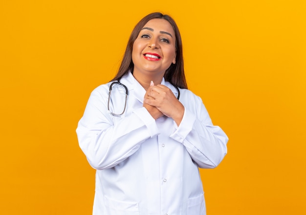 Ärztin mittleren Alters im weißen Kittel mit Stethoskop, die nach vorne schaut, glücklich und positiv lächelnd, fröhlich die Hände zusammenhaltend über orangefarbener Wand haltend