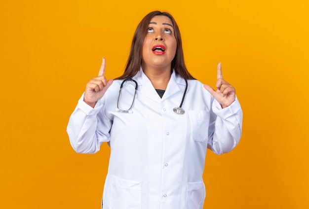 Ärztin mittleren Alters im weißen Kittel mit Stethoskop, die überrascht aufblickt und mit den Zeigefingern nach oben über orangefarbener Wand steht?