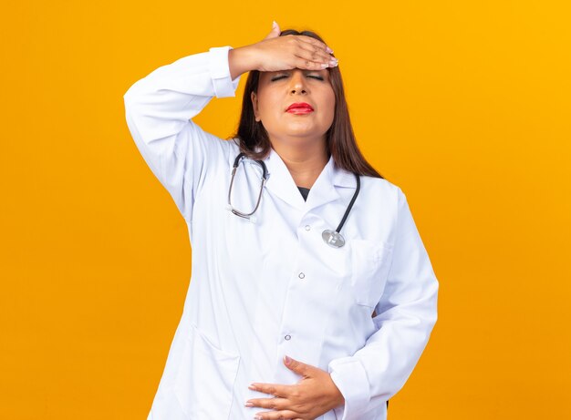 Ärztin mittleren Alters im weißen Kittel mit Stethoskop, die unwohl aussieht und ihre Stirn mit Kopfschmerzen und Fieber berührt