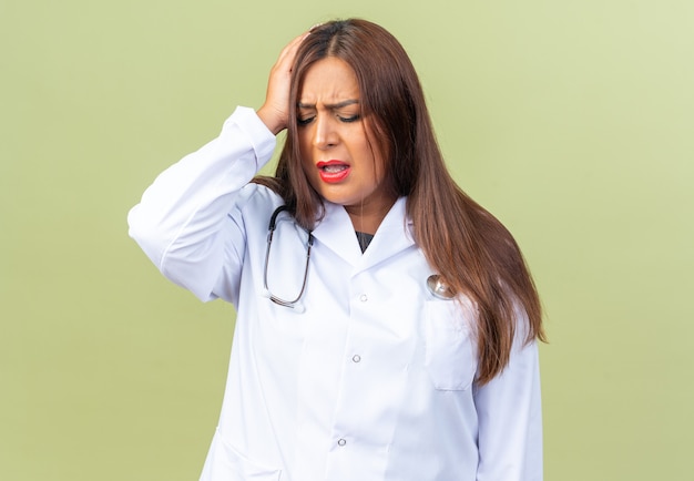 Ärztin mittleren Alters im weißen Kittel mit Stethoskop, die unwohl aussieht und ihren Kopf berührt, der unter starken Kopfschmerzen leidet, die auf Grün stehen