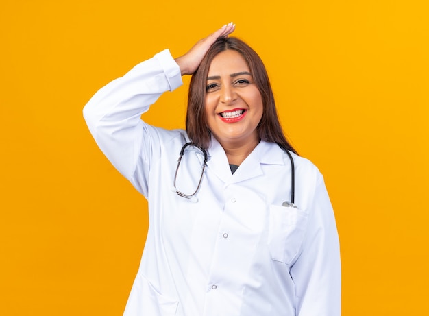 Ärztin mittleren Alters im weißen Kittel mit Stethoskop glücklich und fröhlich, die Hand auf dem Kopf hält