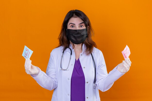 Ärztin mittleren Alters mit weißem Mantel in schwarzer Gesichtsschutzmaske und mit Stethoskop, das Erinnerungspapiere mit Ja- und Nein-Worten hält, die die Kamera betrachten, überrascht über Orange stehend