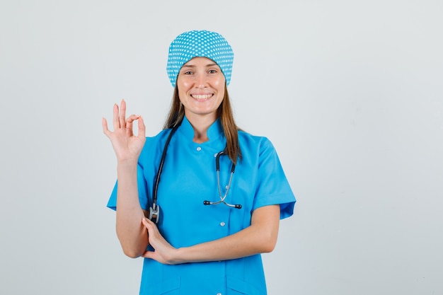 Ärztin zeigt ok Geste in blauer Uniform und sieht fröhlich aus. Vorderansicht.