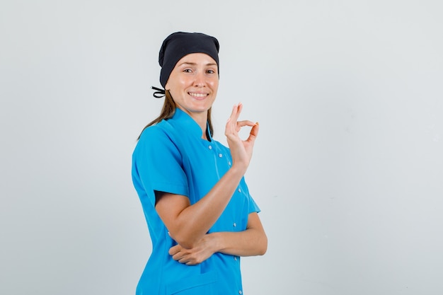 Ärztin zeigt ok Zeichen in blauer Uniform, schwarzem Hut und sieht fröhlich aus. Vorderansicht.