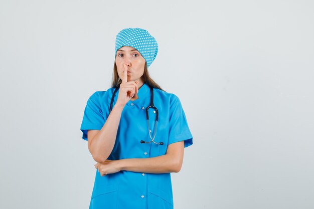 Ärztin zeigt Schweigegeste in blauer Uniform und sieht vorsichtig aus. Vorderansicht.