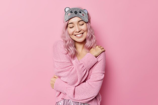 Sanft lächelnde Frau mit gefärbten Haaren umarmt sich und Tagträumen hält die Augen geschlossen, trägt eine Schlafmaske und ein weicher Pullover erinnert an schöne Momente im Leben isoliert über rosa Hintergrund. Self-Care-Konzept