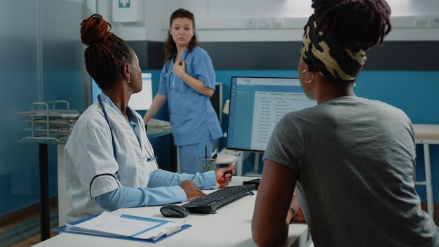 Sanitäter erklärt dem Patienten medizinische Informationen auf dem Computer, während er die Krankenschwester in der Arztpraxis um Hilfe bittet. Arzt, der eine Gesundheitsuntersuchung mit einer Frau unter Verwendung von Monitor und Technologie durchführt