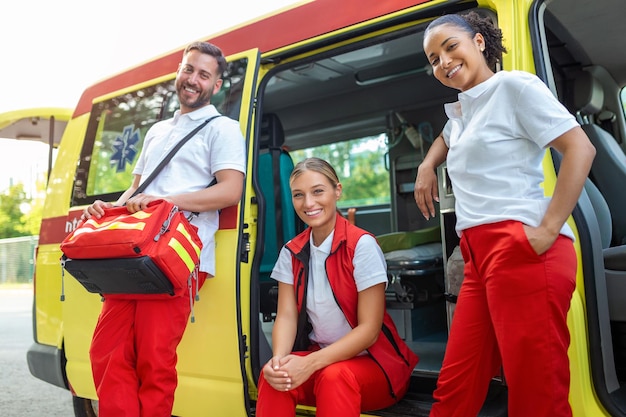 Sanitäter und Arzt stehen auf der Seite des Krankenwagens. Der Arzt trägt einen medizinischen Traumabeutel. Gruppe von drei Sanitätern, die mit einem Lächeln vor dem Krankenwagen stehen