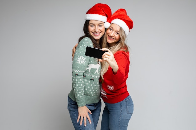Santa Mädchen Freunde in roten Hüten posieren mit Telefon für festliches Selfie auf grauem Hintergrund mit Kopierraum