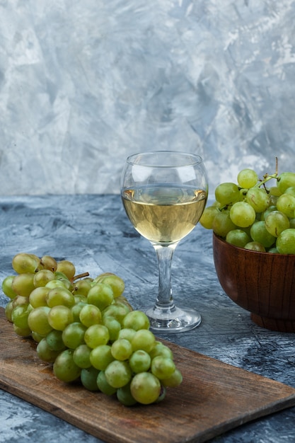 Kostenloses Foto satz glas wein, trauben auf einem schneidebrett und weiße trauben in einer schüssel auf einem dunklen und hellblauen marmorhintergrund. nahansicht.