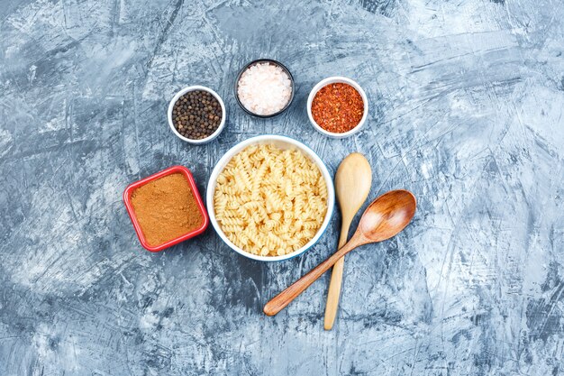 Satz Holzlöffel, Gewürze und Fusilli-Nudeln in einer weißen Schüssel auf einem grauen Gipshintergrund. Draufsicht.