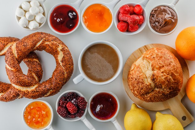 Satz Marmeladen, Himbeere, Zucker, Schokolade in Tassen, türkischer Bagel, Brot, Orange und Zitronen und eine Tasse Kaffee auf einer weißen Oberfläche