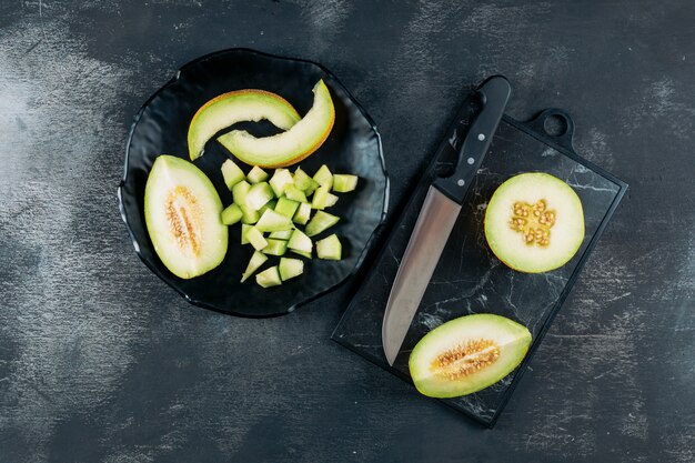 Satz Melone und Messer und geschnittene Melone in einer schwarzen Schüssel auf einem dunklen hölzernen Hintergrund. flach liegen.