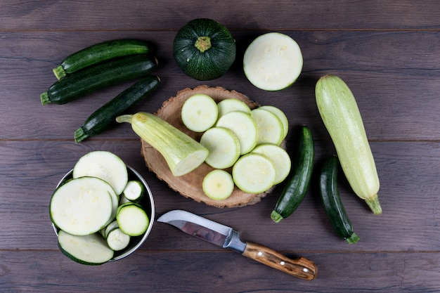 Satz Messer und andere Zucchini in einer Schüssel und herum und geschnittene Zucchini auf einem dunklen Holztisch. flach liegen.