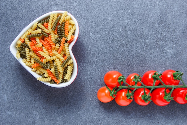 Kostenloses Foto satz von tomaten und bunten makkaroni-nudeln in einer herzförmigen schüssel auf einer grauen oberfläche. draufsicht. platz für text