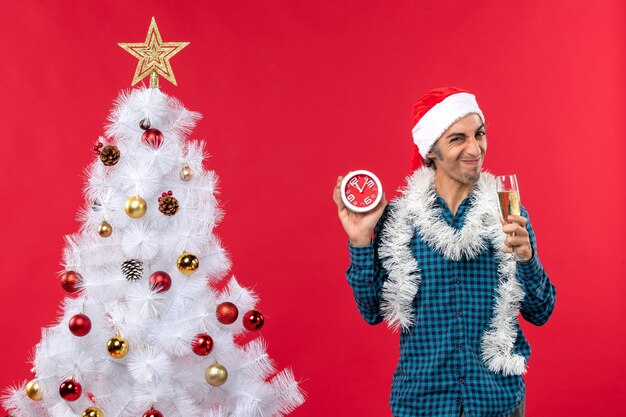 Saurer Gesicht junger Mann mit Weihnachtsmannhut und ein Glas Wein und halten Uhr, die nahe Weihnachtsbaum auf Rot steht