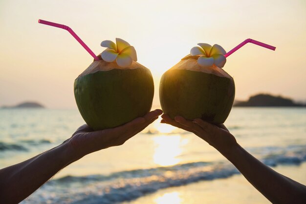 Schattenbild der frischen Kokosnuss in den Paarhänden mit dem Plumeria verziert auf Strand mit Seewelle - Tourist mit Sonnenferienkonzept der frischen Frucht und des Meersands