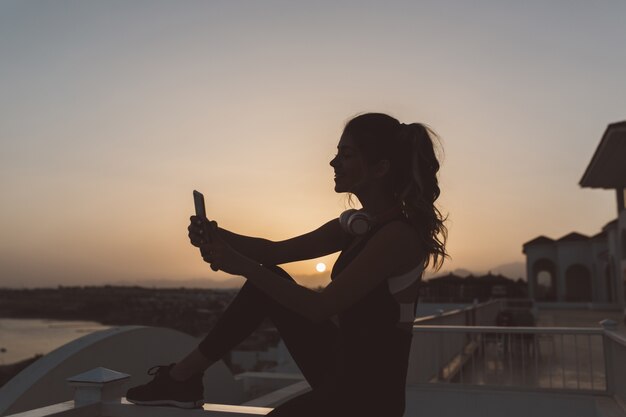 Schattenbild der jungen attraktiven Frau in der Sportbekleidung, die selfie am Meer auf Sonnenuntergang macht. Positivität ausdrücken, aktiven Lebensstil, genießen, fröhliche Stimmung.