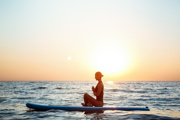 Schattenbild der schönen Frau, die Yoga auf Surfbrett bei Sonnenaufgang praktiziert.