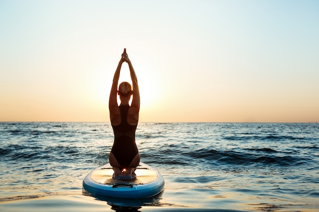 Schattenbild der schönen Frau, die Yoga auf Surfbrett bei Sonnenaufgang praktiziert.