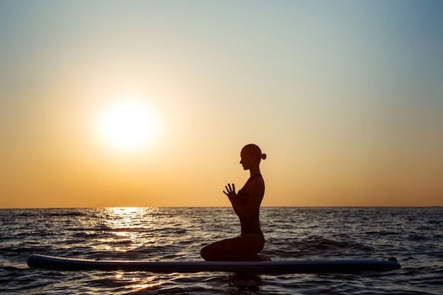 Schattenbild der schönen Frau, die Yoga auf Surfbrett bei Sonnenaufgang praktiziert.