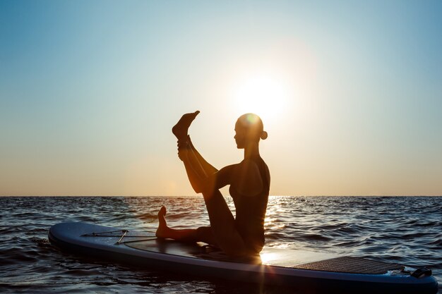 Schattenbild der schönen Frau, die Yoga auf Surfbrett bei Sonnenaufgang praktiziert.