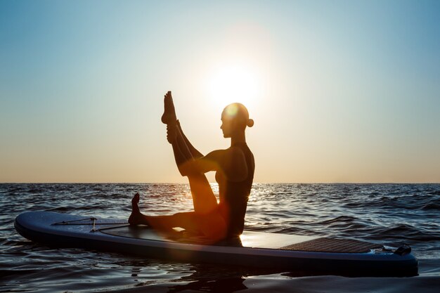 Schattenbild der schönen Frau, die Yoga auf Surfbrett bei Sonnenaufgang praktiziert.