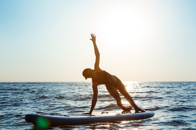 Schattenbild der schönen Frau, die Yoga auf Surfbrett bei Sonnenaufgang praktiziert.