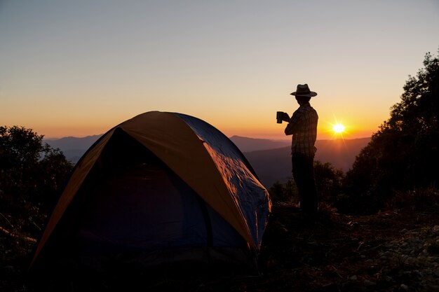 Schattenbild des glücklichen Mannes mit dem Halten des Kaffeetasseaufenthaltes nahe Zelt um Berge