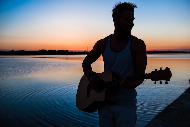 Schattenbild des jungen gutaussehenden Mannes, der Gitarre am Meer während des Sonnenaufgangs spielt