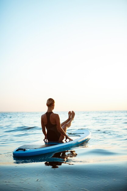 Schattenbild des jungen schönen Mädchens, das Yoga auf Surfbrett im Meer bei Sonnenaufgang praktiziert.