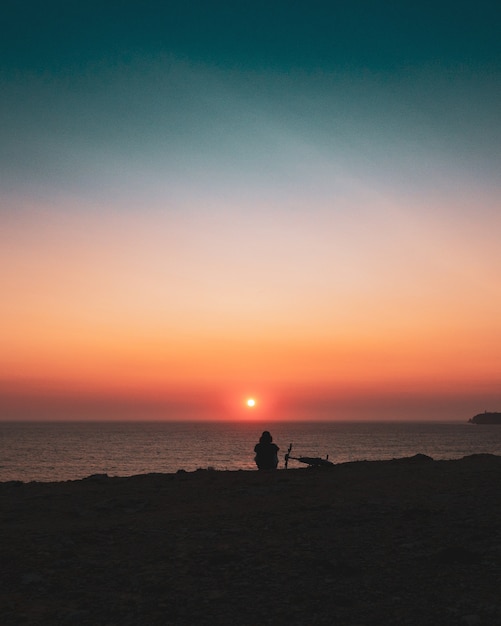 Schattenbild einer Person, die am Ufer während des Sonnenuntergangs sitzt