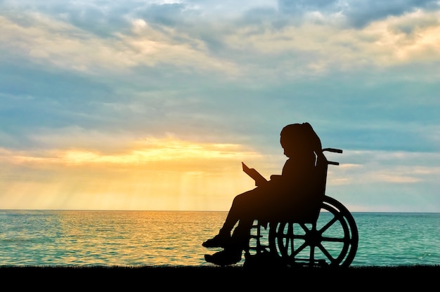 Schattenbild eines behinderten kindermädchens, das in einem rollstuhl sitzt und ein buch auf einem seesonnenunterganghintergrund liest. konzeptuelles bild des lebens von kindern mit behinderungen