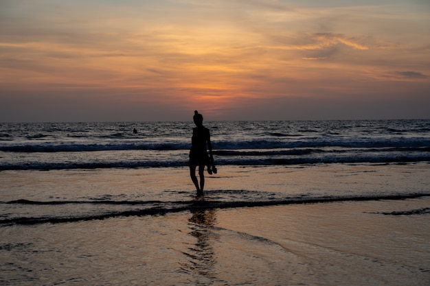 Schattenbild eines Mädchens, das in der Hand auf das Wasser auf einem Strand mit ihren Schuhen geht, während die Sonne untergeht