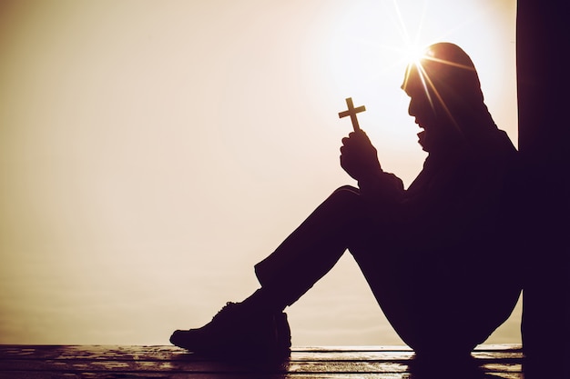 Kostenloses Foto schattenbild eines mannes, der in der hand mit einem kreuz bei sonnenaufgang betet.