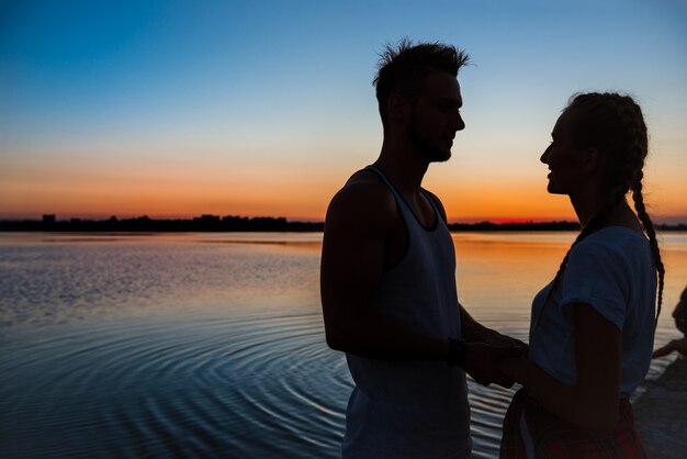 Schattenbilder des jungen schönen Paares, das sich am Sonnenaufgang nahe See erfreut