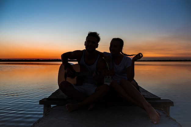 Schattenbilder des jungen schönen Paares, das sich am Sonnenaufgang nahe See erfreut